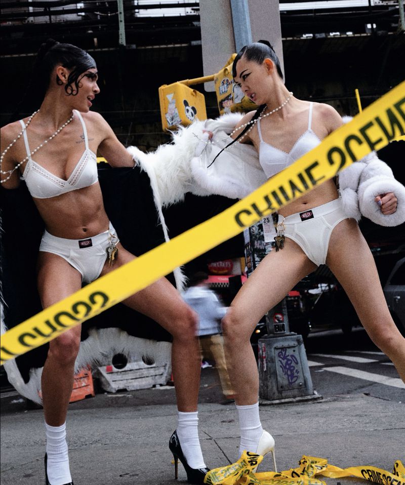 Two adult female models in white lingerie and socks, posed in an outdoor urban setting, surrounded by yellow 'Crime Scene' tape.
