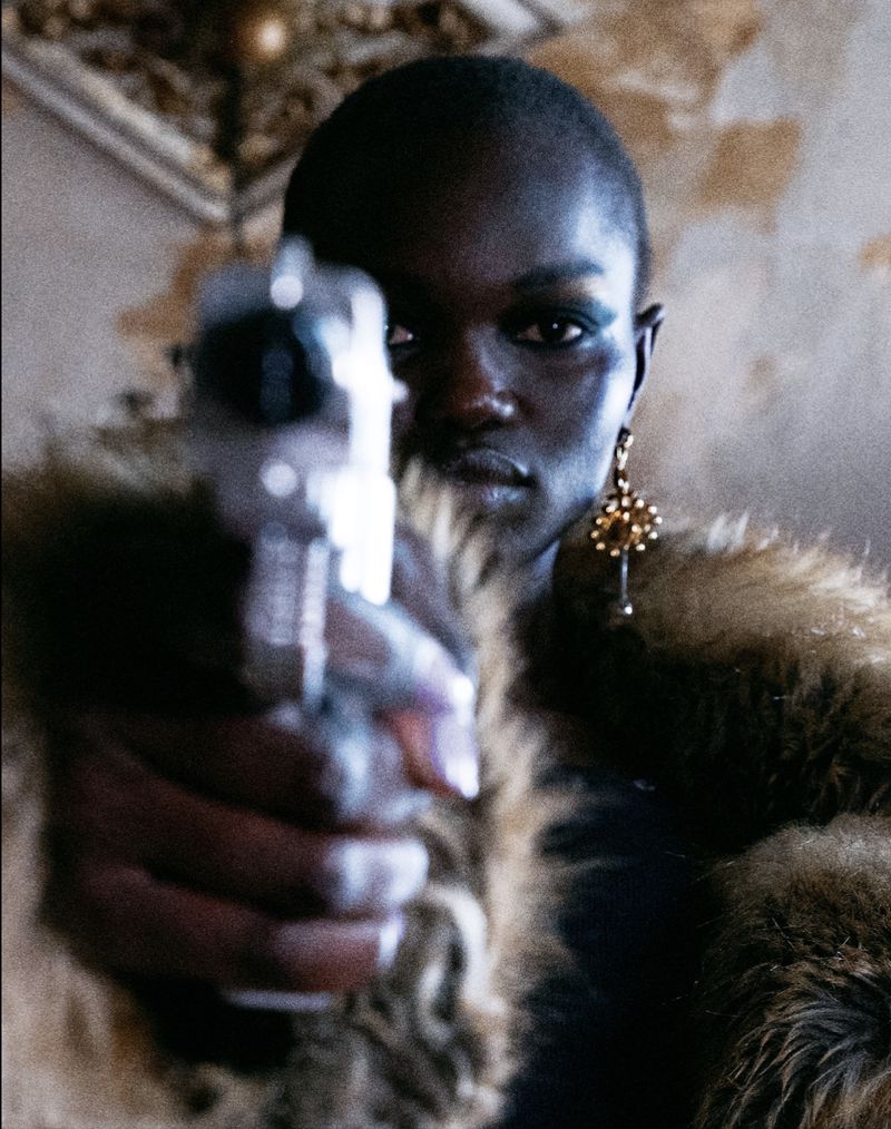 Close-up of a dark-skinned woman pointing a silver handgun directly at the viewer, wearing gold earrings and a fur coat.
