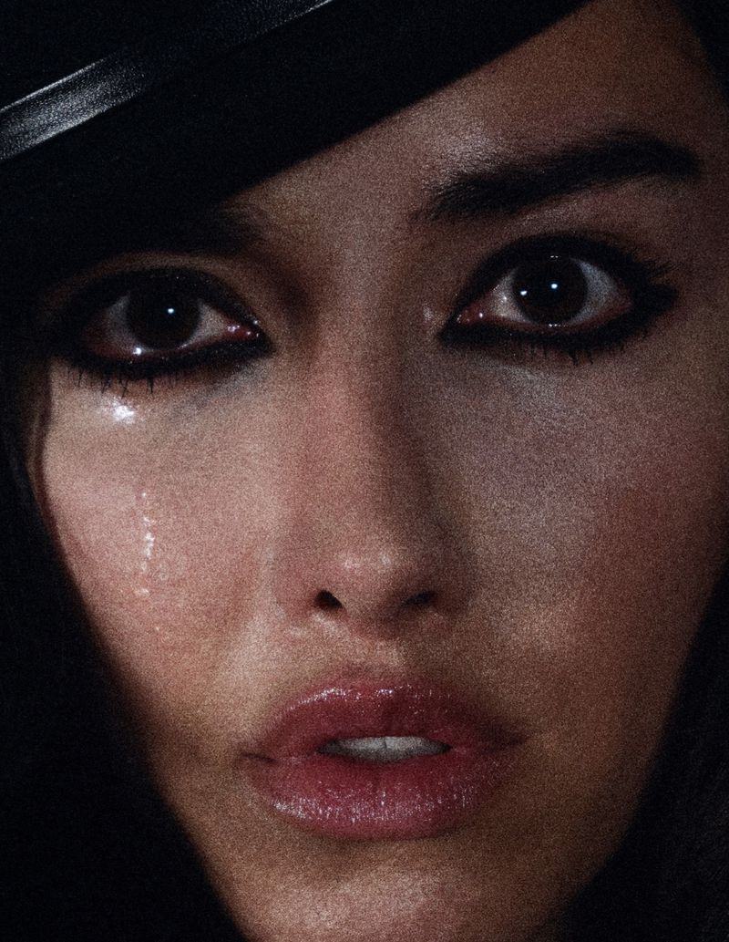 Close-up portrait of a woman's face with tears, featuring dramatic eye makeup and glossy lips in a dark, moody setting.