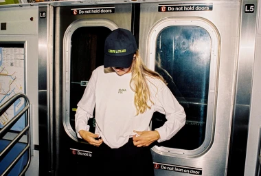 Person wearing a navy embroidered baseball cap and white MoMA PS1 long-sleeve t-shirt inside a subway train car.