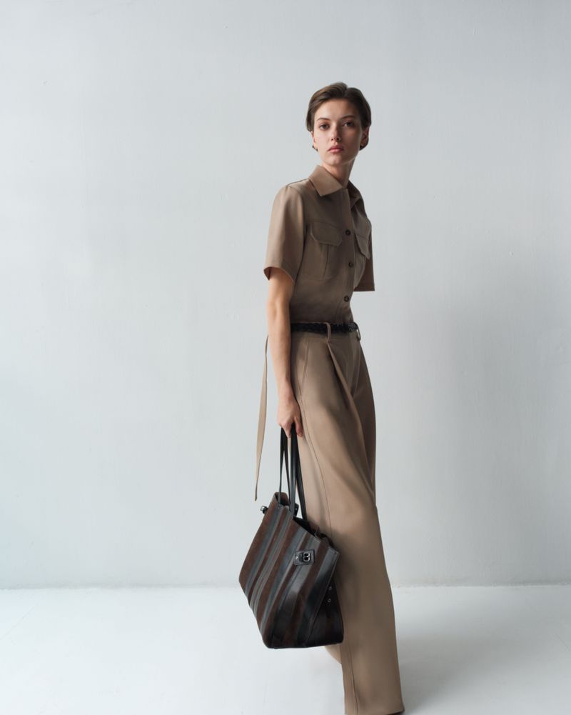Valerie Scherzinger in a tan tailored short-sleeve shirt and wide-leg trousers holding a striped leather tote against a white backdrop.