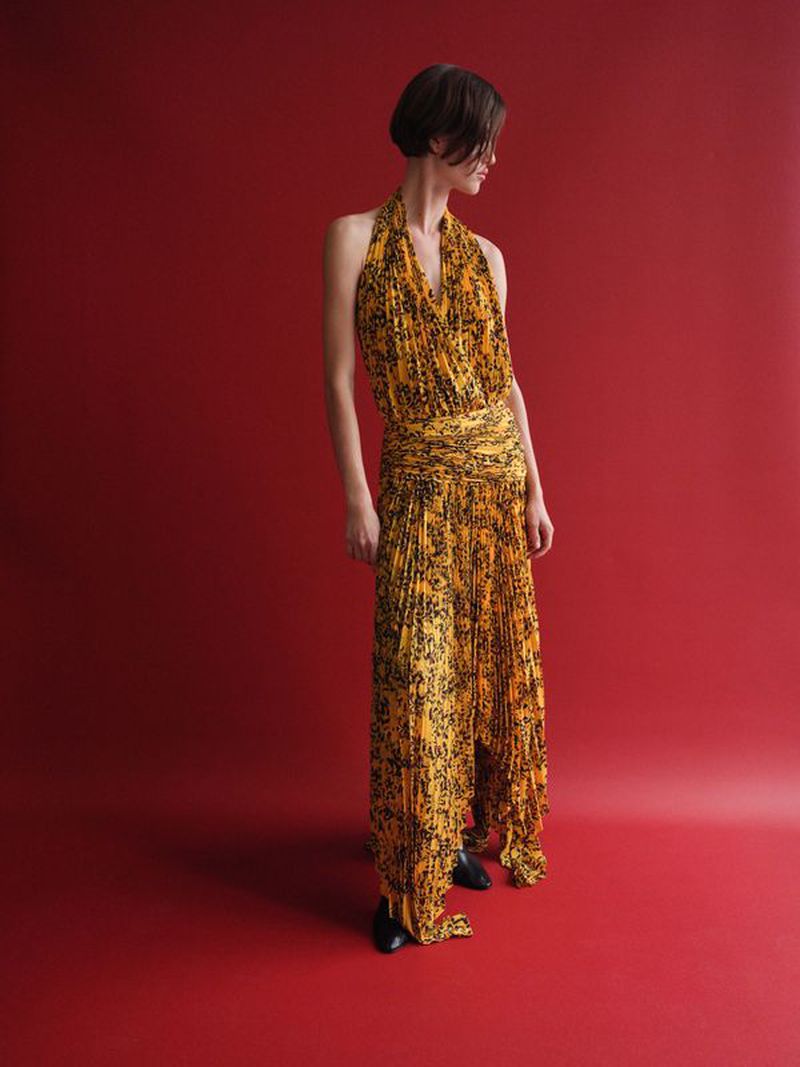 Model Valerie Scherzinger in a yellow and black pleated halter dress standing against a rich red studio background.