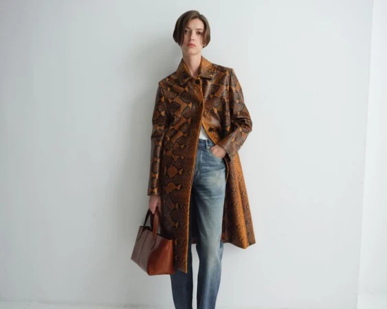 Valerie Scherzinger wearing a brown snakeskin print coat and blue denim jeans while holding a brown leather tote bag against a white wall.