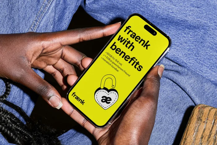 Black hands holding a smartphone showing the fraenk with benefits campaign graphic on a bright yellow screen over blue denim.