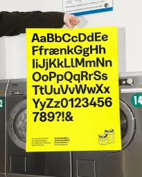 Hand holding a bright yellow typography poster displaying the alphabet in front of stainless steel washing machines in a laundromat.