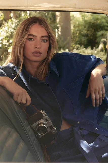 Woman in blue jacket holding a camera inside a car, posing with a neutral expression, trees visible in the background, medium shot.