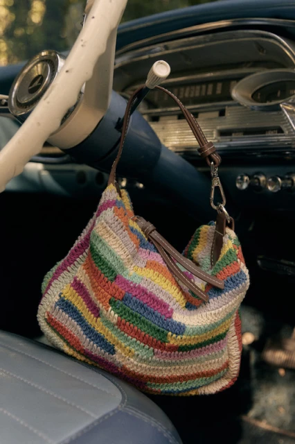 A multi-colored crocheted hobo bag is suspended from the steering wheel of a classic blue car, casting shadows inside the vehicle.