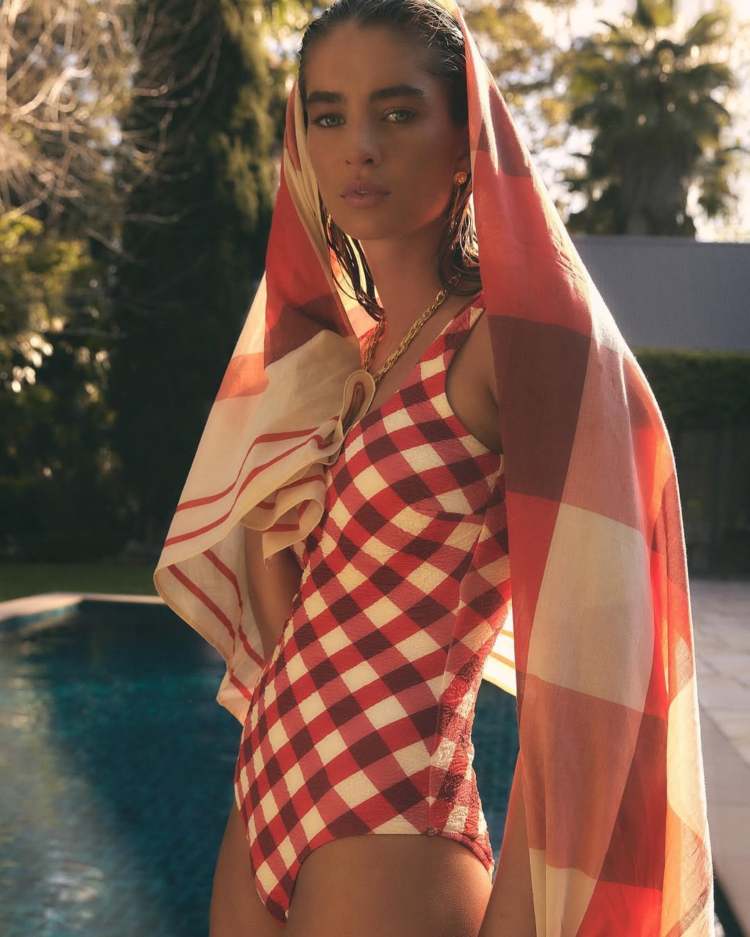 Woman in red and white checkered swimsuit poses near pool with towel draped over head in outdoor setting, looking at camera.