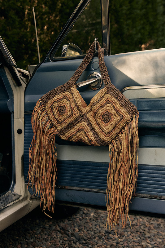 A crocheted brown and beige fringe bag hangs on the open door of a blue vintage car, with green trees blurred in the background.