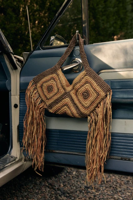 A crocheted brown and beige fringe bag hangs on the open door of a blue vintage car, with green trees blurred in the background.