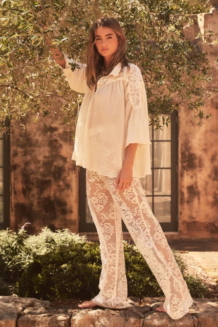A woman poses outdoors wearing a white lace top and bell-bottom pants, with a brick building and tree visible in the background.