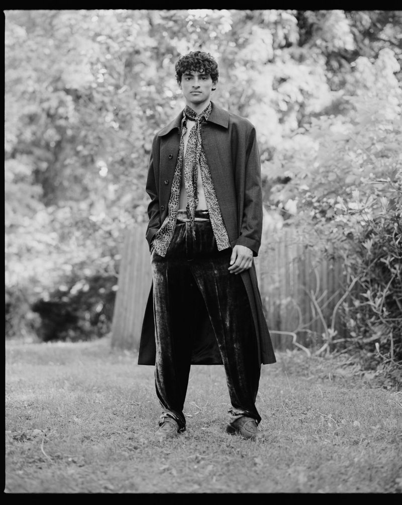 Monochrome portrait of a slender adult male in a coat, patterned shirt, and velvet trousers standing in a grassy outdoor setting.