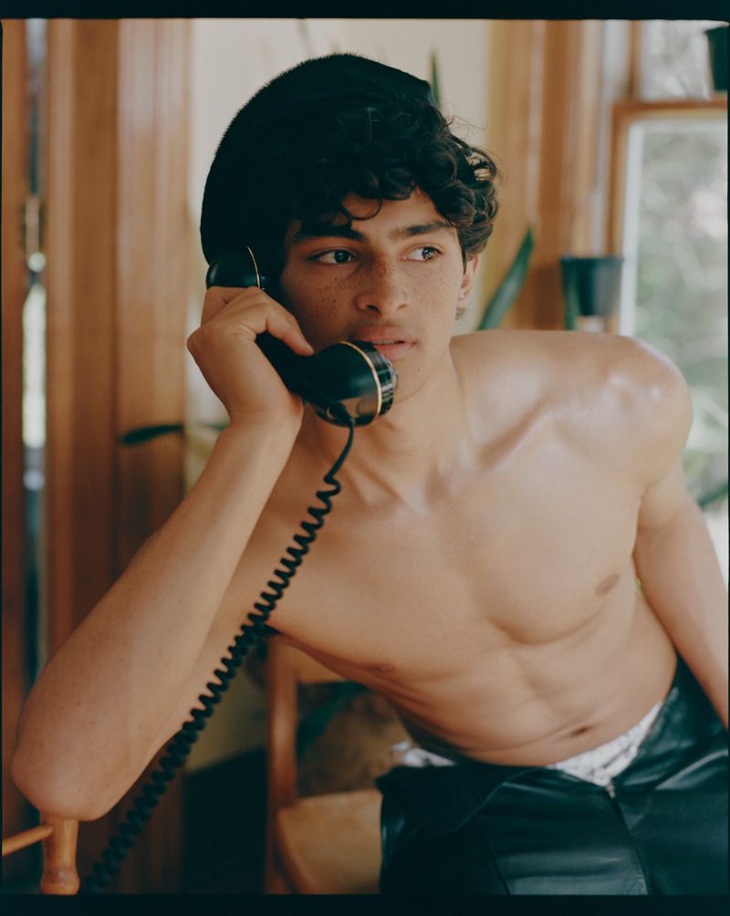 Young man with dark hair and no shirt, wearing a black cap, is talking on a retro telephone in an indoor setting with natural light.