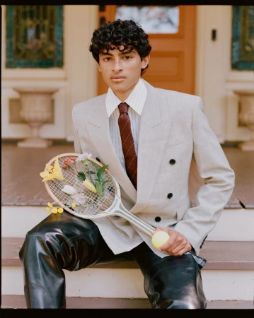 Young man in a blazer sitting on stairs with tennis racket filled with flowers and a tennis ball in hand, with a building in the background.