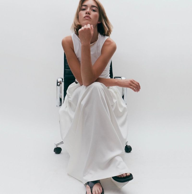 Model wearing white top and maxi skirt seated on office chair against white seamless background