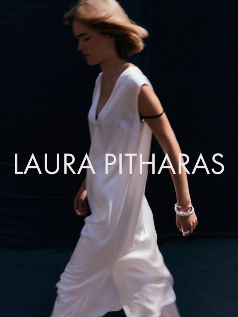 Model walking in a sleeveless white V-neck dress with silver jewelry and black armband against a dark backdrop.