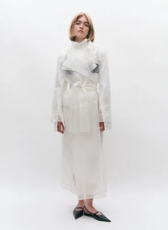 A woman posing in a sheer white belted trench coat and black pointed flat shoes against a plain white studio background.