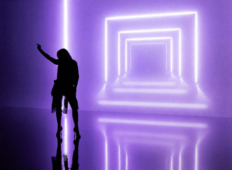 Silhouette of Madonna posing in high heels in front of glowing purple neon rectangular frames on a reflective studio floor.