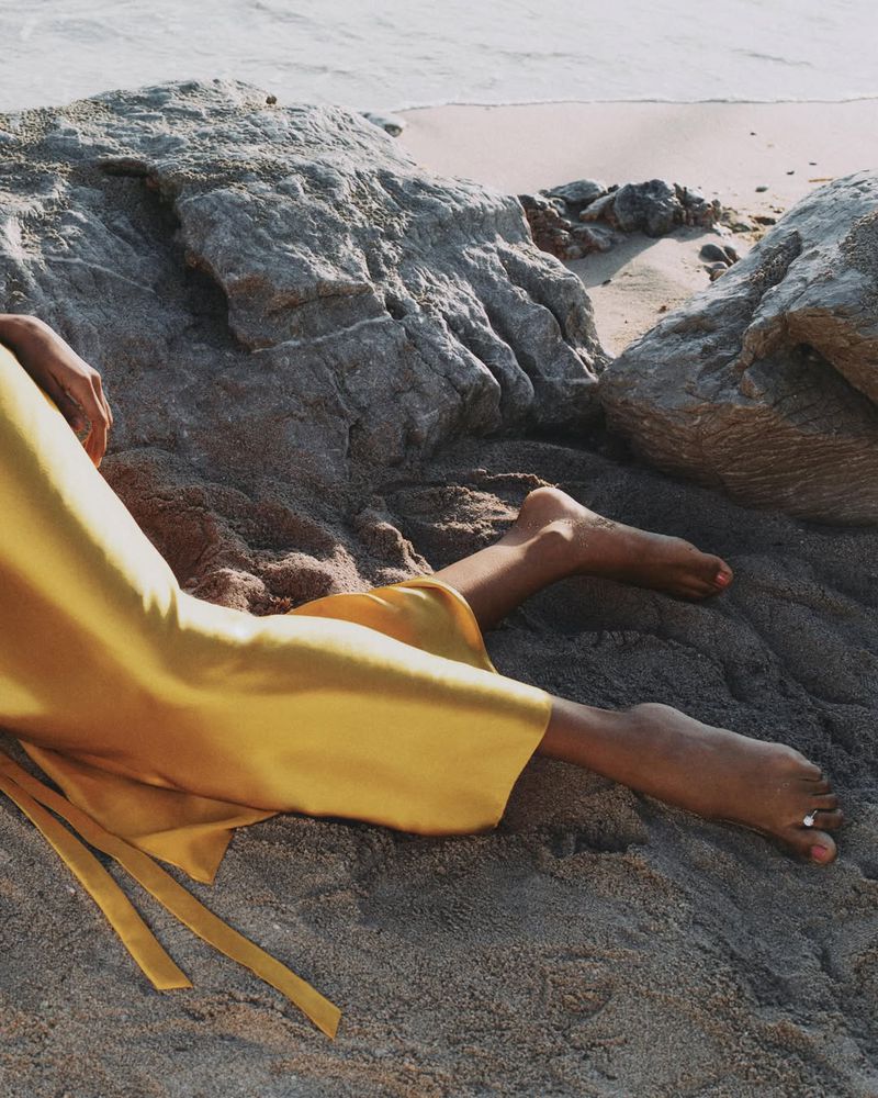 Close-up of a model in a vibrant yellow satin dress lying barefoot on a sandy beach next to coastal rocks.