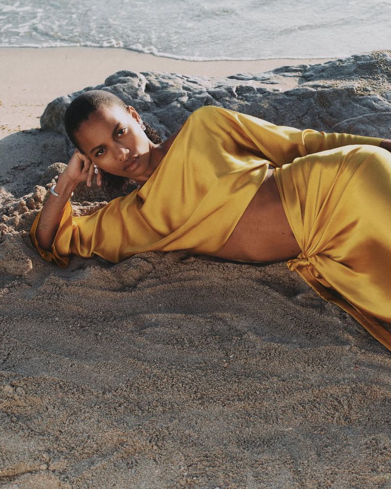 Model lying on a sandy beach wearing a draped golden yellow satin crop top and matching gathered skirt.