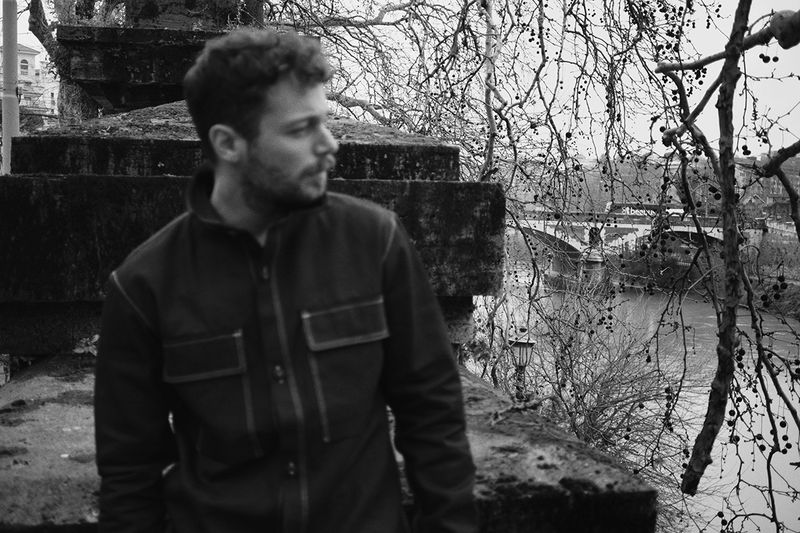 Monochrome portrait of a man in a jacket standing near a stone structure, with a bridge and trees visible in the background.