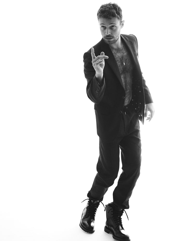 Monochrome portrait of a man in a suit jacket, pants, and combat boots, captured in a full shot against a clean white background.