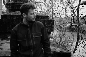 Portrait of Ludovico Tersigni wearing a dark jacket outdoors near a river bridge for Next Gen by Simone Paccini