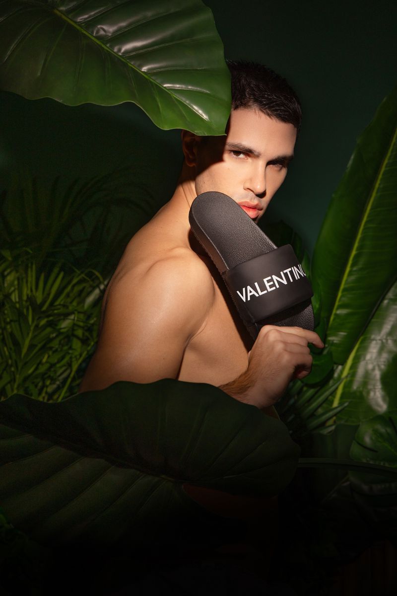Shirtless male model posing behind tropical green leaves while holding a black slide sandal featuring white Valentino branding.