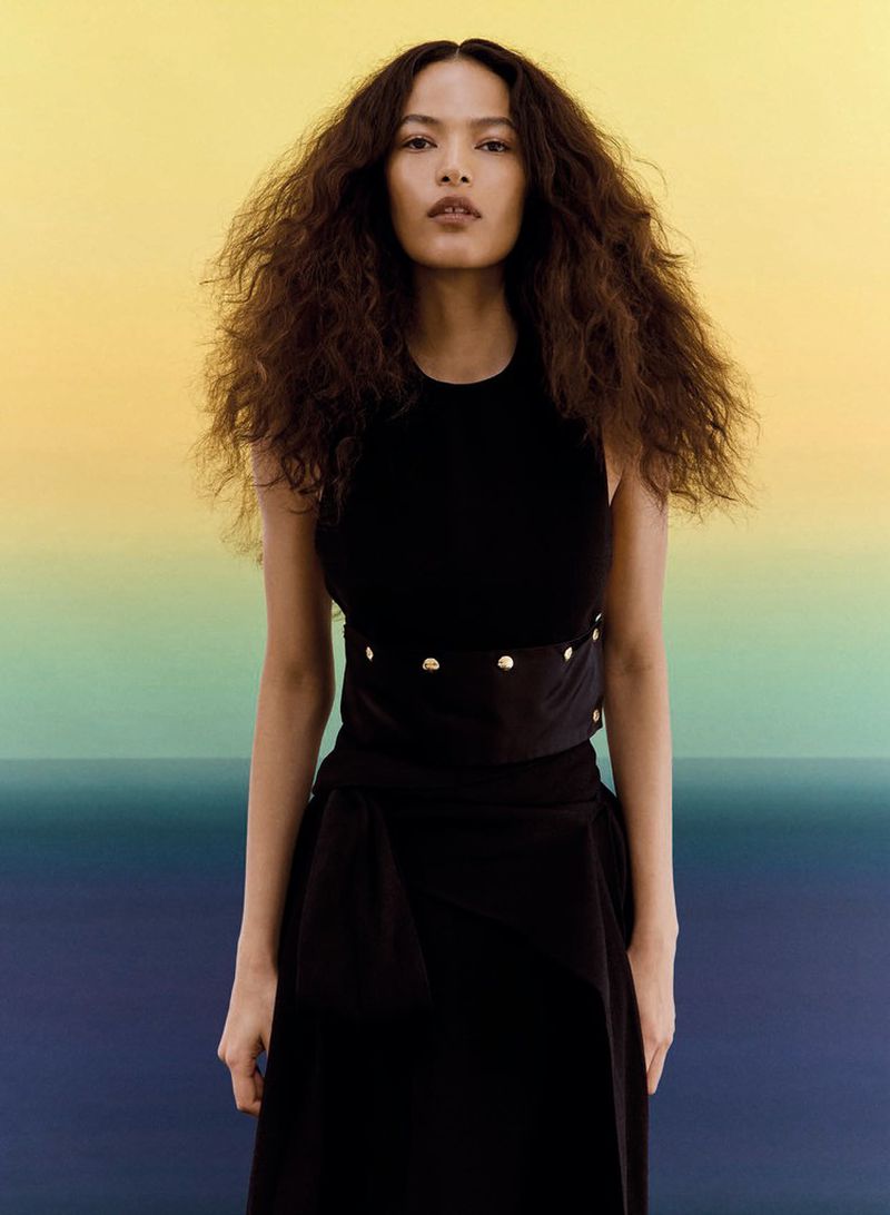 Model Luana Guimarães in a sleeveless black dress with waist detail against a yellow, green, and blue gradient background.