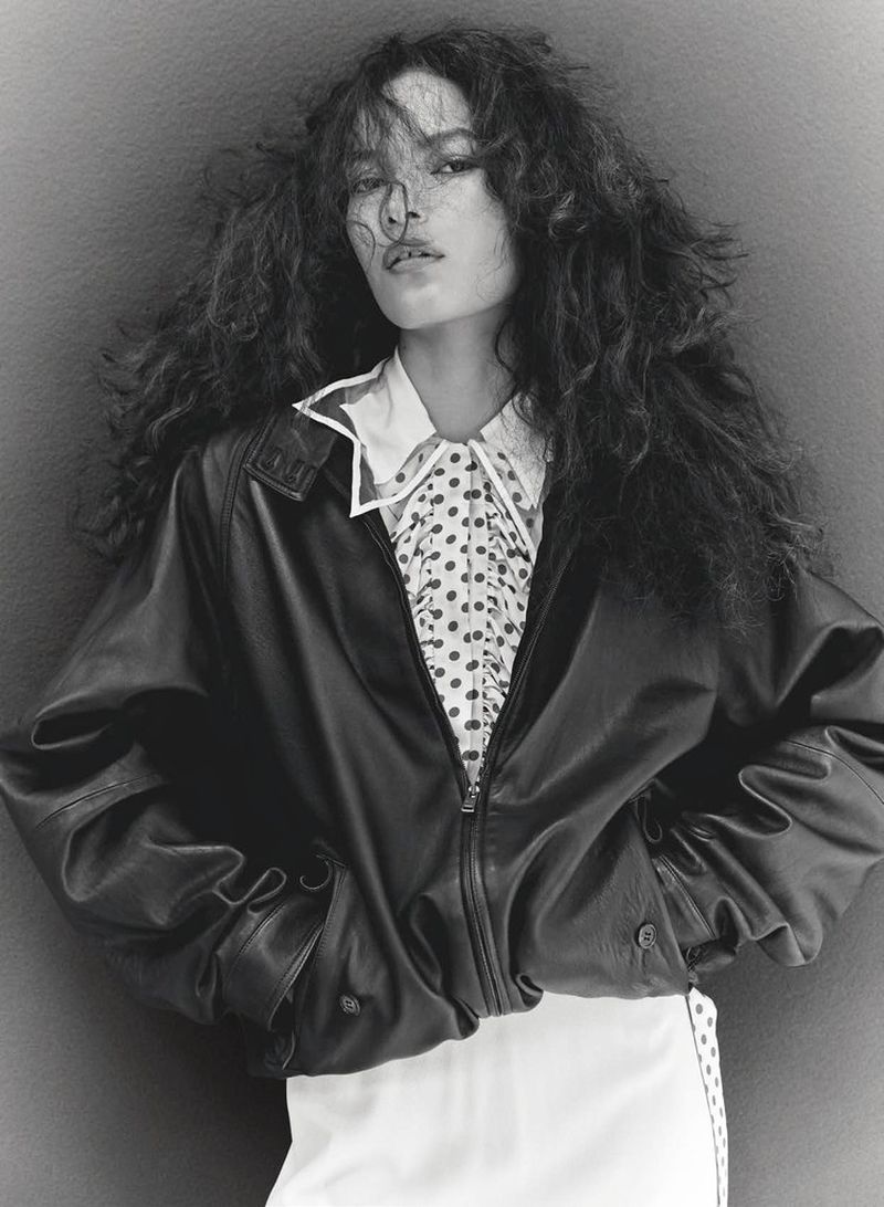 Model Luana Guimarães in an oversized black leather jacket and polka-dot collared shirt against a neutral studio backdrop.
