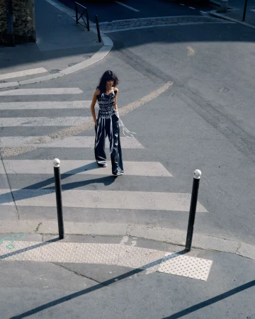 High-angle view of a woman walking across an asphalt crosswalk wearing wide dark trousers and a fringe-embellished sleeveless top.