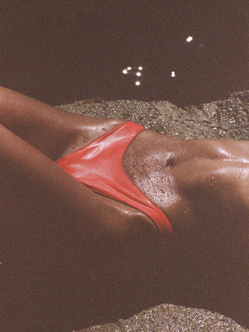 Close-up of a model with wet skin lying on coastal rocks wearing bright red bikini bottoms.
