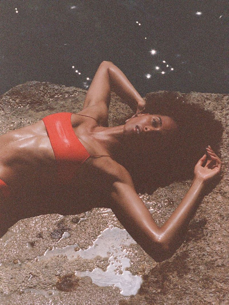 Model Cindy Bruna reclines on coastal rocks wearing an orange-red bikini top next to sparkling ocean water.