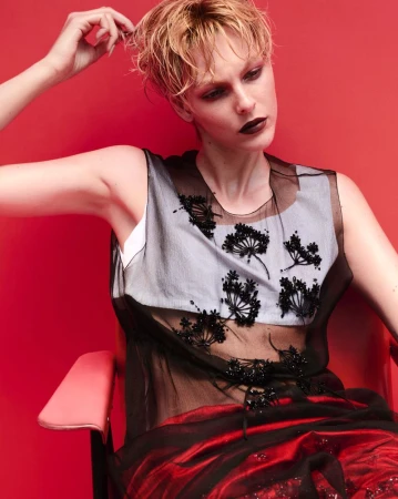 Vittoria Ceretti seated against a bold red backdrop wearing a sheer black floral embroidered top and red skirt.
