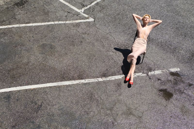 Vittoria Ceretti sits on a chair with arms behind her head on an asphalt parking lot marked with white painted lines.