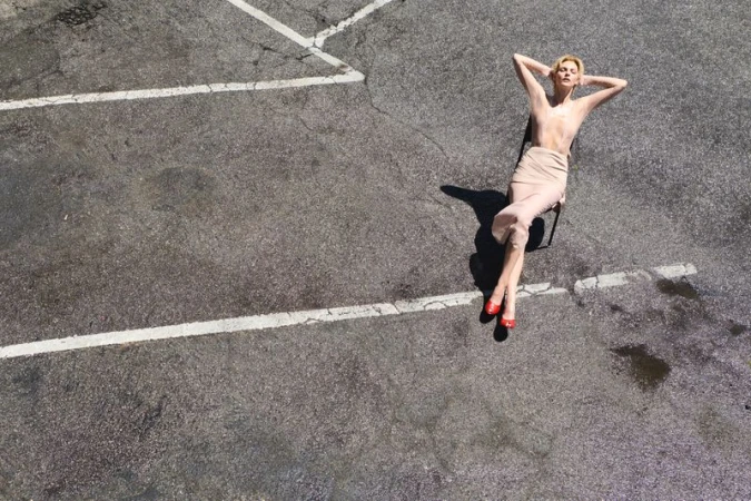 Vittoria Ceretti sits on a chair with arms behind her head on an asphalt parking lot marked with white painted lines.