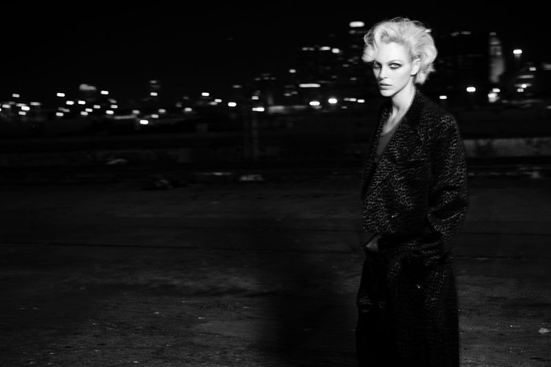 Vittoria Ceretti with short blonde hair wearing a dark patterned coat at night against a blurred city skyline backdrop.