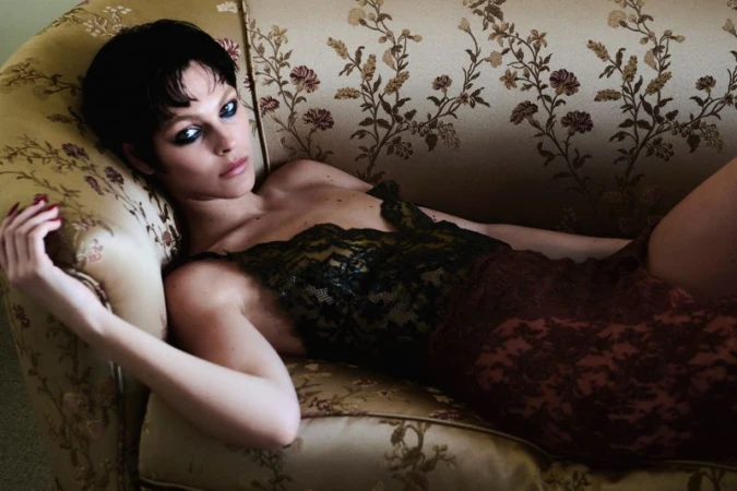 Model Vittoria Ceretti reclining on a vintage floral upholstered sofa wearing a dark lace dress with smoky eye makeup.