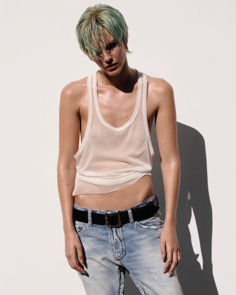 Model Vittoria Ceretti with dyed green hair wearing a sheer white tank top and denim jeans against a white background.