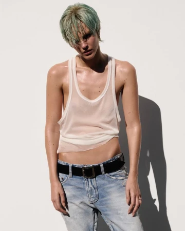 Model Vittoria Ceretti with dyed green hair wearing a sheer white tank top and denim jeans against a white background.