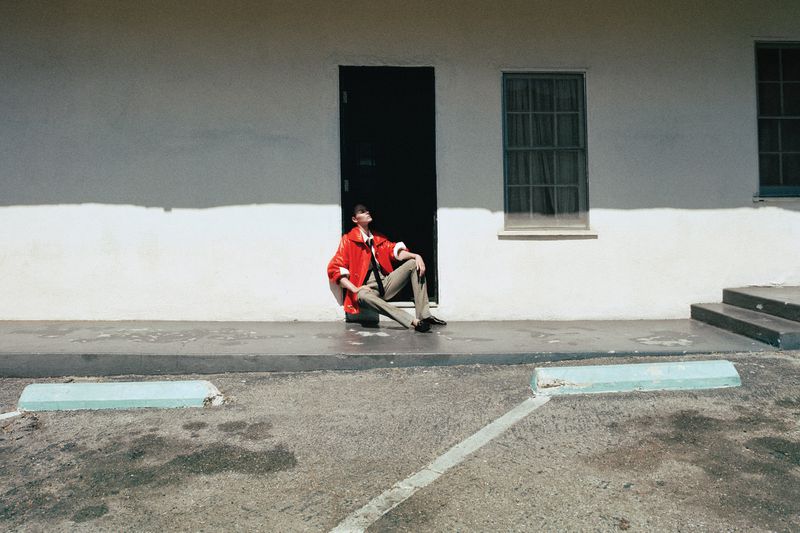 Vittoria Ceretti seated in front of a black doorway wearing a bright red jacket and beige trousers in bright sunlight.