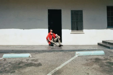 Vittoria Ceretti seated in front of a black doorway wearing a bright red jacket and beige trousers in bright sunlight.