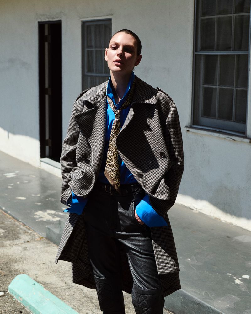Model Vittoria Ceretti in an oversized houndstooth trench coat and blue shirt standing outdoors in bright sunlight against a white wall.