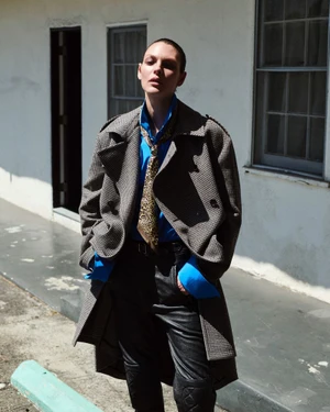 Model Vittoria Ceretti in an oversized houndstooth trench coat and blue shirt standing outdoors in bright sunlight against a white wall.
