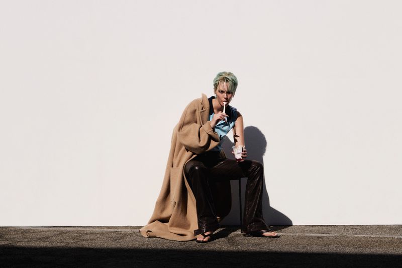 Vittoria Ceretti in a camel coat, blue top, and leather pants seated against a bright white wall sipping an iced drink.