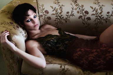 Model Vittoria Ceretti reclining on a vintage floral upholstered sofa wearing a dark lace dress with smoky eye makeup.