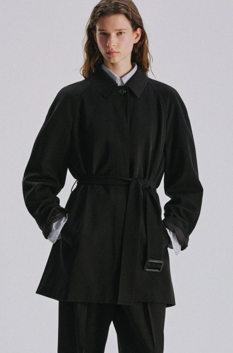 Model Sandra Murray wearing a black belted coat and tailored trousers against a pale grey studio background.