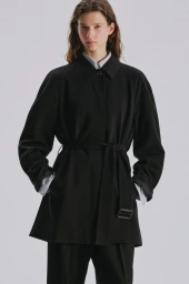 Model Sandra Murray wearing a black belted coat and tailored trousers against a pale grey studio background.