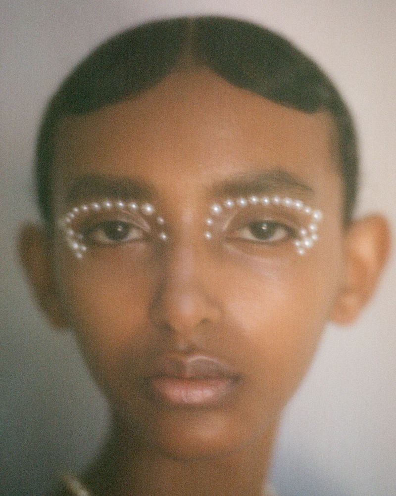 Close-up portrait of a young adult woman with pearls as makeup around her eyes, against a light grey studio background.