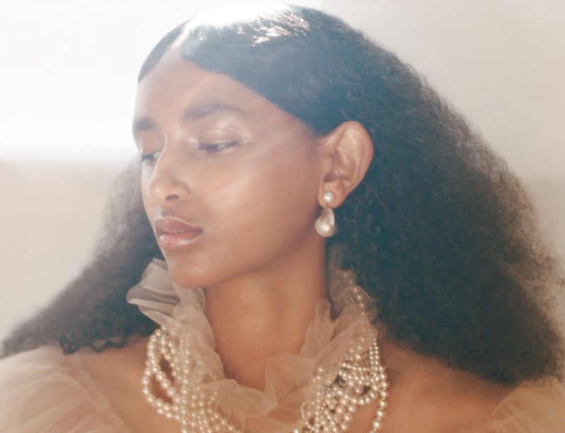 Elegant portrait of a Black woman with pearl jewelry and soft lighting. She is wearing a ruffled top with her head angled and eyes closed.